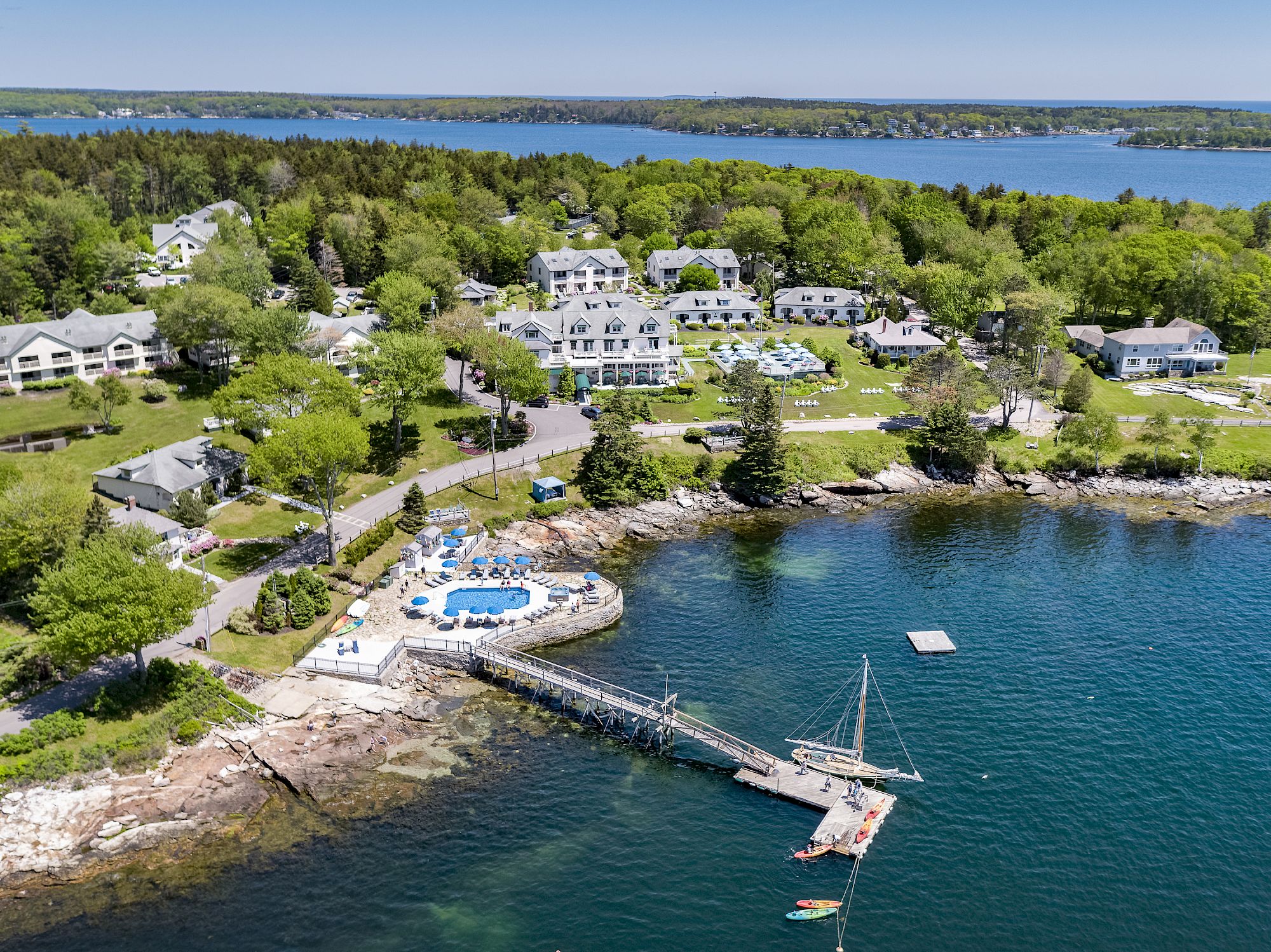 Aerial view of a coastal resort with a dock, boats, a pool, and several buildings surrounded by trees near the water’s edge, ending the sentence.