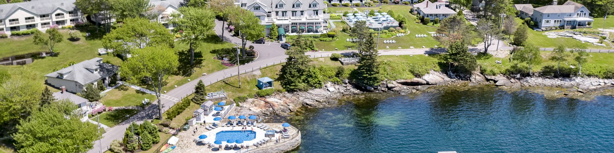 An aerial view of a coastal resort with multiple buildings, a swimming pool, a pier, and a sailboat, surrounded by lush greenery and water.