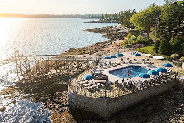 A coastal resort with a pool area, sun loungers, and umbrellas, next to a rocky shore and calm water, with a clear sky overhead.