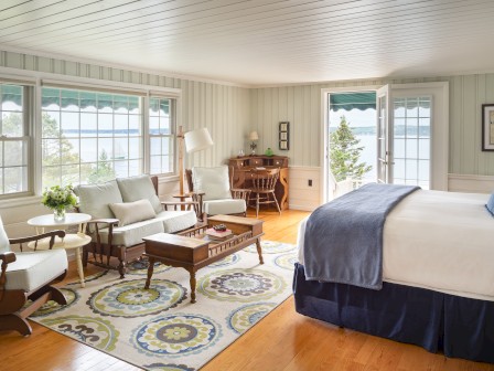 A bright and spacious bedroom with large windows, white furniture, a patterned rug, a bed, and a desk with a view of a scenic landscape.