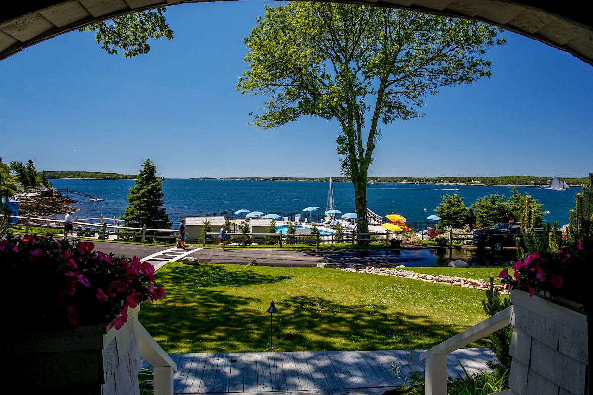 The image shows a serene lakeside scene with a lush green lawn, boats docked, and a clear blue sky, viewed from a porch with flowers ending the sentence.