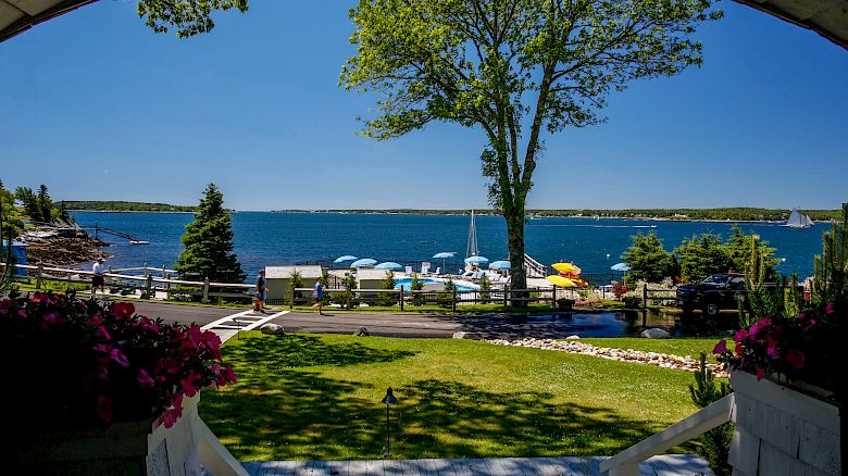 The image shows a serene lakeside scene with a lush green lawn, boats docked, and a clear blue sky, viewed from a porch with flowers ending the sentence.