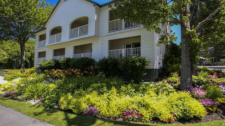 A white two-story house with balconies, surrounded by lush greenery, colorful flowers, and a tree on a sunny day.