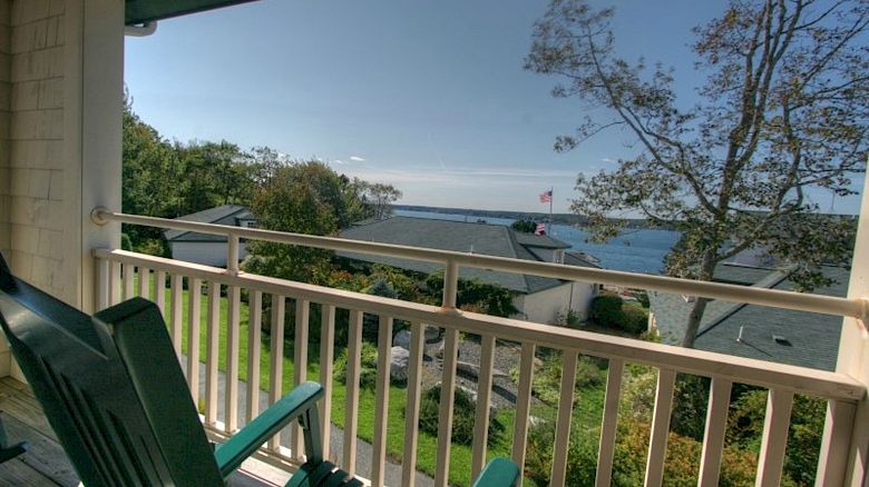 Two green chairs on a balcony overlook a scenic view of houses, trees, and a body of water under a clear blue sky.