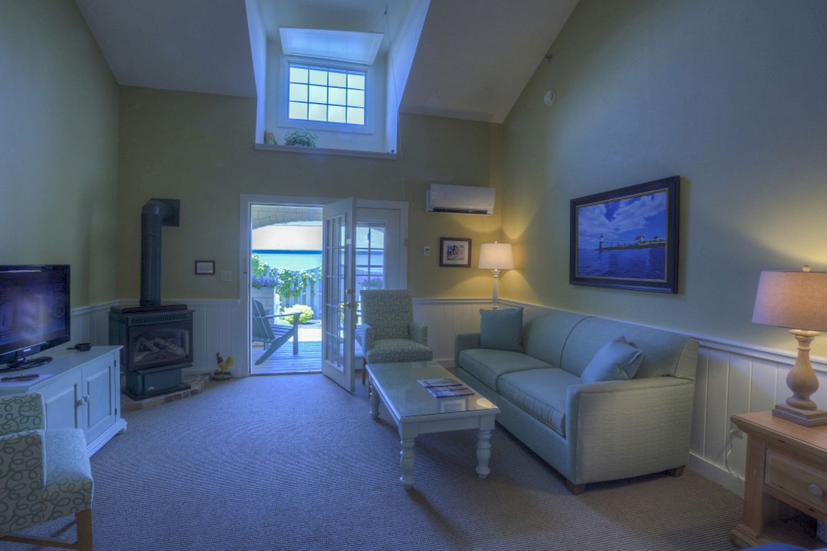 A cozy living room with a sofa, coffee table, lamp, painting, TV, wood stove, and a windowed door opening to a patio with an ocean view.