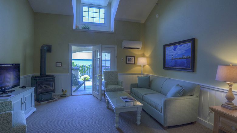 A cozy living room with a sofa, coffee table, lamp, painting, TV, wood stove, and a windowed door opening to a patio with an ocean view.