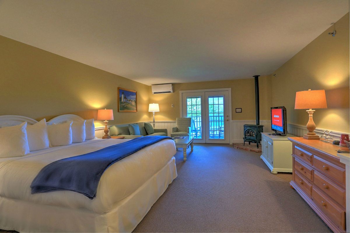 A spacious hotel room with a large bed, TV, dresser, seating area, and lamps, leading to a balcony with French doors; warmly lit and cozy.