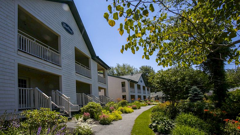 A picturesque path with lush landscaping leads to a row of charming, white, two-story buildings under a clear blue sky.