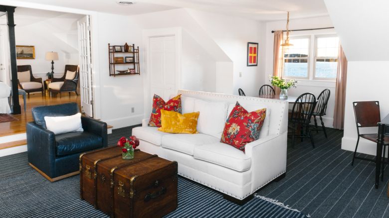 A cozy living room with a white couch, colorful pillows, a trunk coffee table, and a blue armchair near a dining area with dark wood furniture.