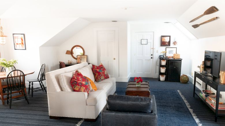 A cozy living space with a white sofa, colorful cushions, a small dining area, and wall art. A TV stand and storage units are also present.