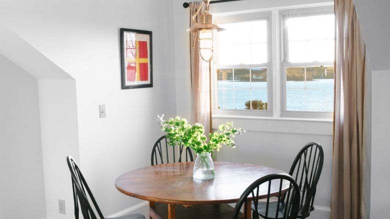 A cozy dining room with a round table, four chairs, wall art, and a window with beige curtains. A vase with flowers sits on the table.