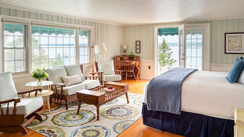 A cozy bedroom with large windows, a bed, a sitting area, a desk, and a view of a lake or ocean through French doors ending the sentence.