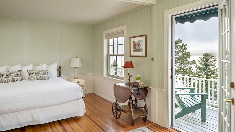 A bedroom with a white bed, wooden floors, a small desk, a lamp, and a rug. An open door leads to a balcony with a chair and forest view.