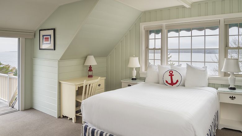 A cozy bedroom with a white bed, nautical-themed pillow, desk, two lamps, a window with an ocean view, and light green walls with slanted ceiling.