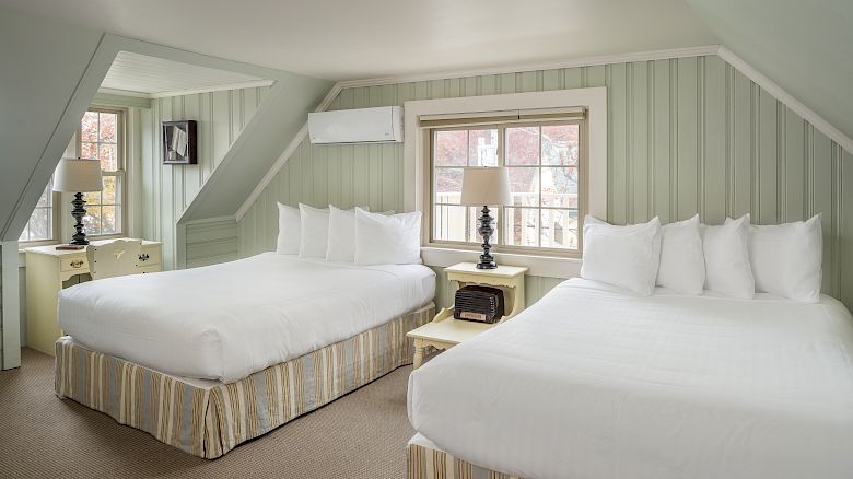 A cozy room with two double beds, nightstands with lamps, a window, a dresser with a TV, pillows, an air conditioning unit, and light green walls.