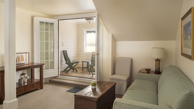 This image shows a cozy living room with a sofa, a wooden chest, a table, and a doorway leading to a small, enclosed porch.