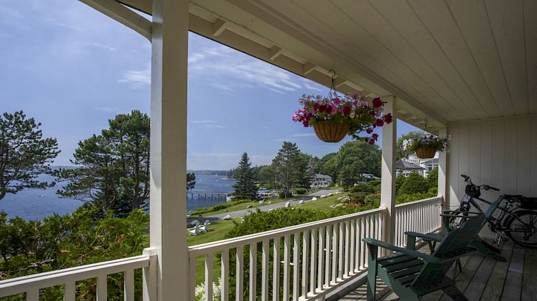 A porch overlooks a scenic view with trees, water, and houses in the distance; chairs and a bicycle are on the porch.