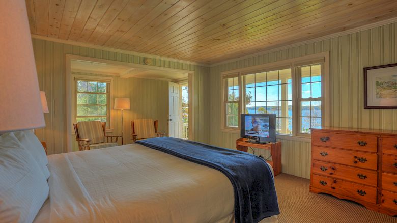 A cozy bedroom with a wooden ceiling, a bed with a blue throw, a TV on a dresser, and two chairs by the window.