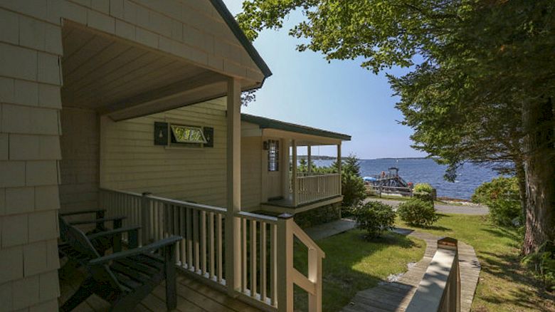 A cozy lakeside cabin with a porch, surrounded by trees, overlooking a dock and the serene water under the bright sky.