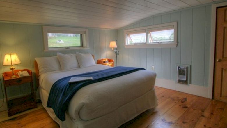 A cozy bedroom with wooden floors, a neatly made bed, blue blanket, bedside tables, lamps, and small windows letting in natural light.