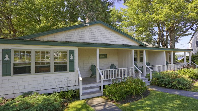 A single-story house with a front porch, green-trimmed shutters, surrounded by greenery with trees in the background and a well-kept lawn ending the sentence.