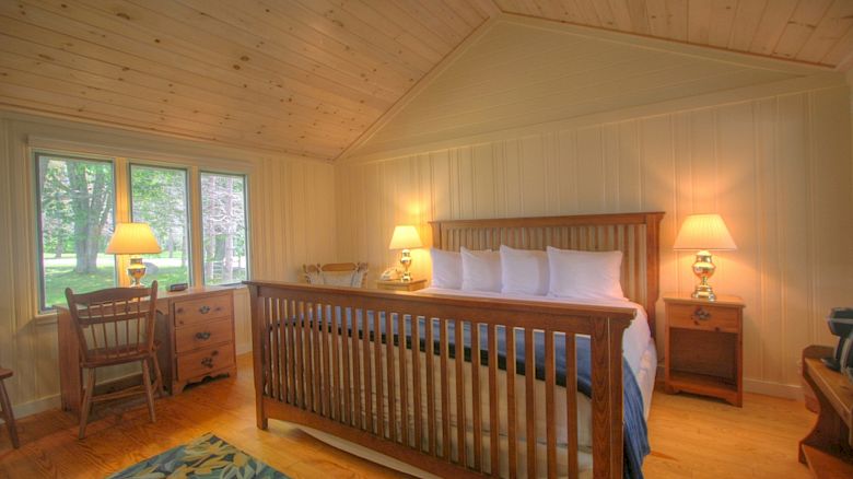 A cozy bedroom with a wooden ceiling and walls, featuring a large bed, two nightstands with lamps, a desk, chair, and window.