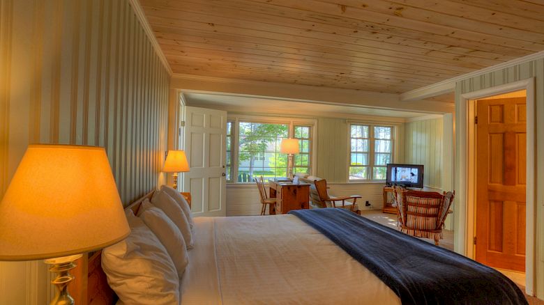 The image shows a cozy bedroom with wooden ceiling and walls, a large bed, lamps, and a seating area with a table, chairs, and a TV.