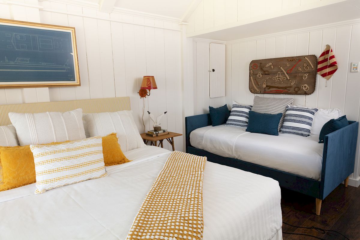 A cozy bedroom featuring a double bed and a daybed, decorated with white and yellow bedding, blue pillows, and nautical-themed wall art and accessories.