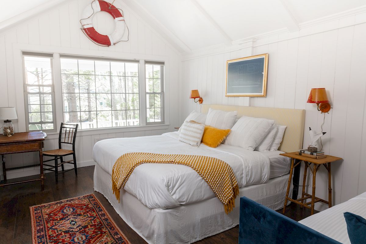 A cozy bedroom with a large bed, white walls, a window, and a desk. Decor includes a life preserver ring, a rug, and framed art.