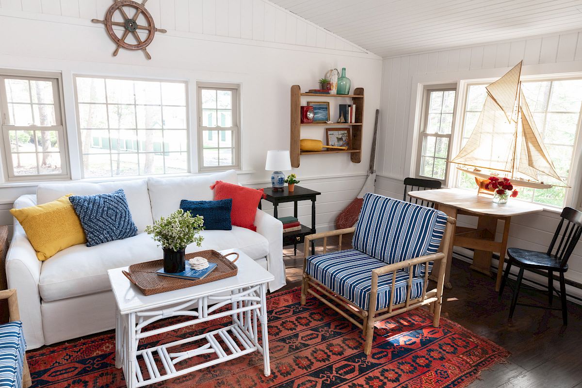 A cozy living room with nautical decor features a white sofa, striped chairs, a sailboat model, a red rug, and a bookshelf against white walls.