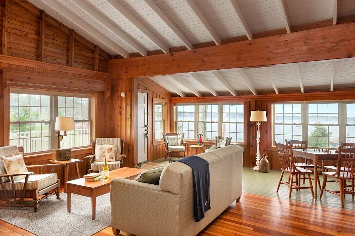 A cozy living room with wooden walls, furniture, large windows, and a view of a lake; includes a sofa, chairs, lamps, and a dining table.