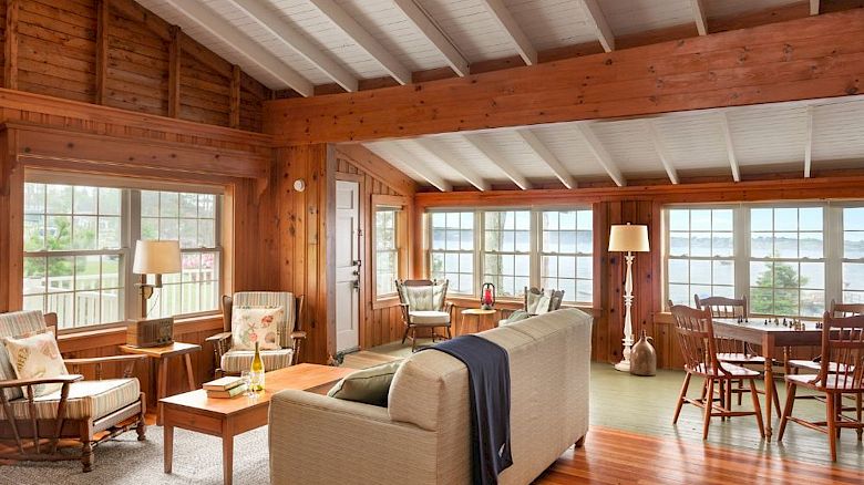 A cozy living room with wooden walls, furniture, large windows, and a view of a lake; includes a sofa, chairs, lamps, and a dining table.