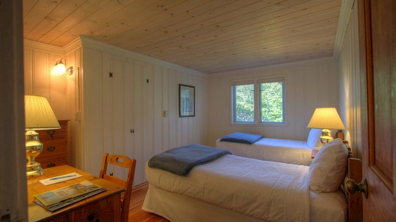 The image shows a cozy bedroom with two single beds, a desk, a chair, lamps on nightstands, a wooden ceiling, and windows letting in natural light.