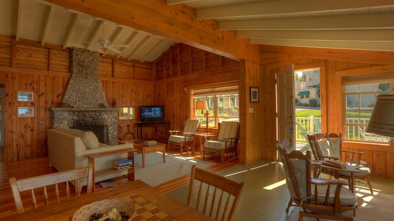 A cozy wooden living room with a stone fireplace, seating area, TV, dining table, and windows letting in sunlight plus a view outside.