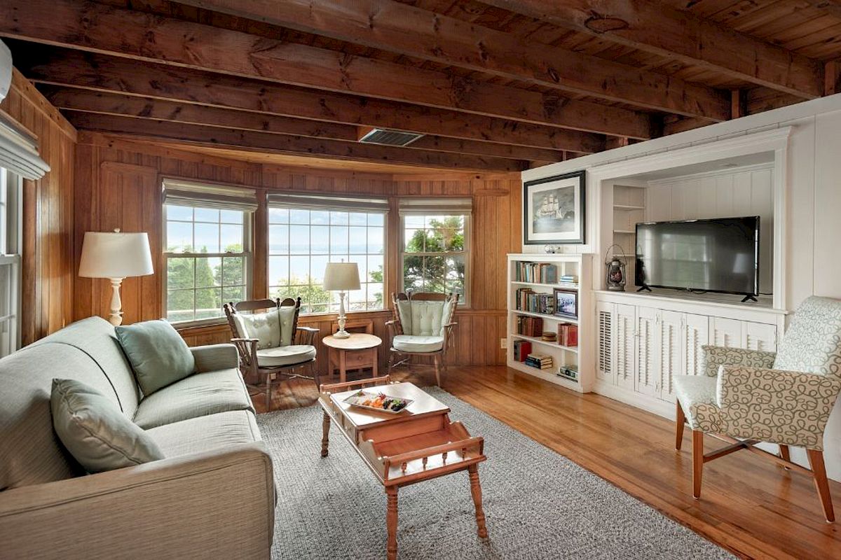 A cozy living room with wood-paneling, large windows, multiple chairs, a sofa, a TV, and a coffee table with a tray of snacks ends the sentence.