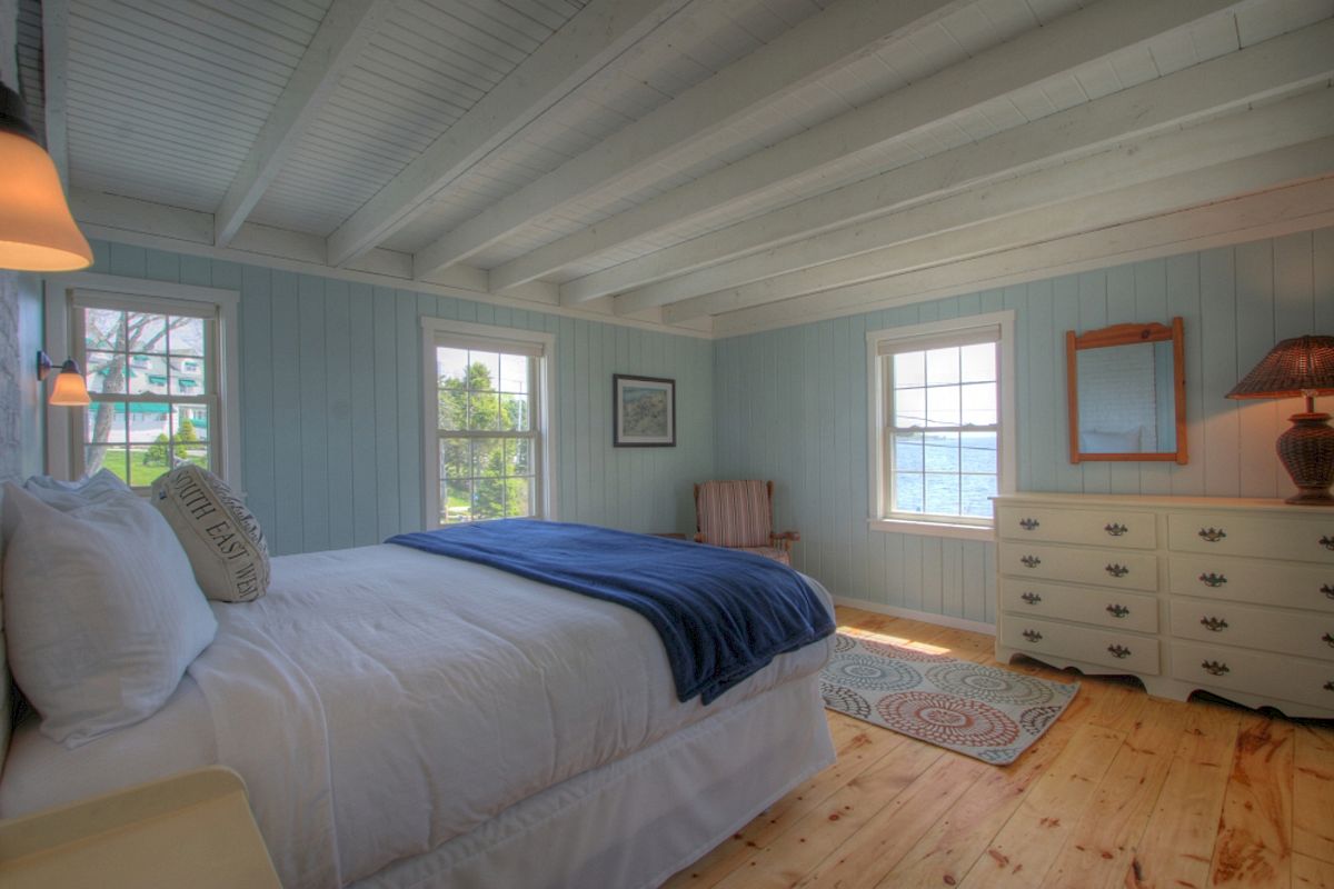 A cozy bedroom with a bed, blue blanket, pillows, dresser, mirror, chair, rug, and windows offering views of a house and trees outside.