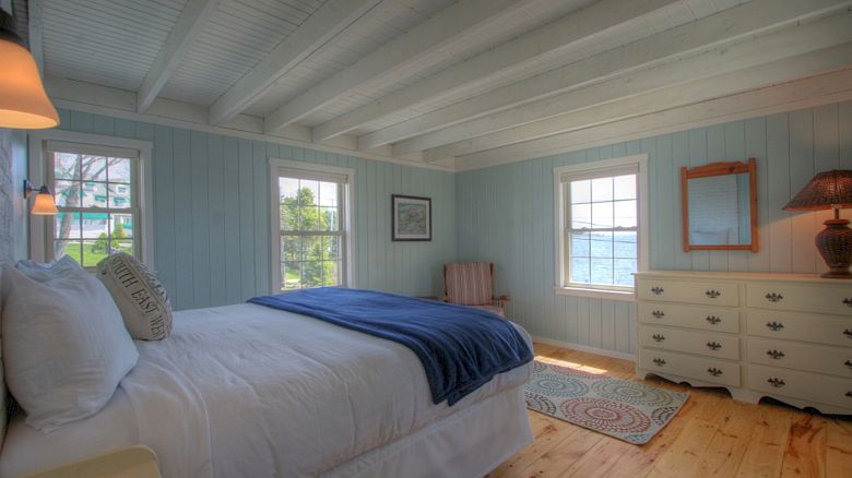 A cozy bedroom with a bed, blue blanket, pillows, dresser, mirror, chair, rug, and windows offering views of a house and trees outside.