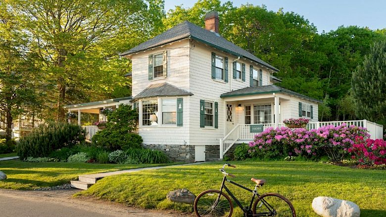 A charming two-story house surrounded by greenery and flowers, with a vintage bicycle parked on the grass beside the road, under a clear sky.