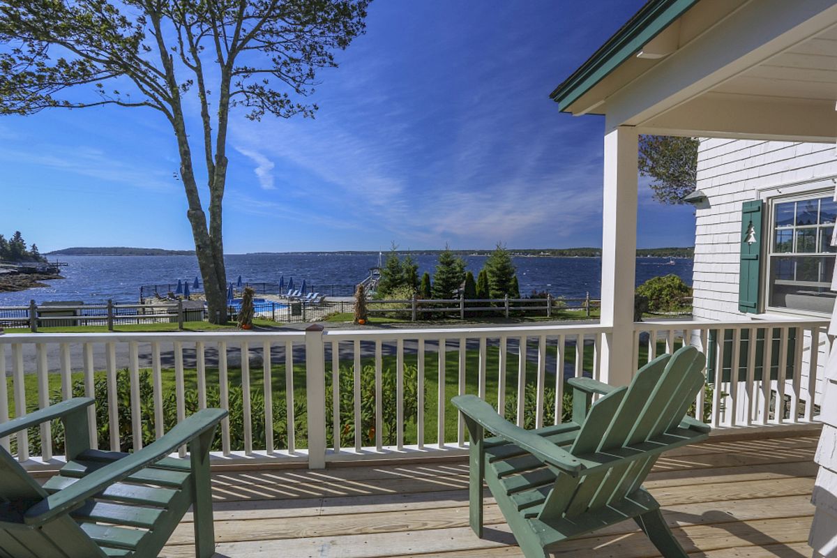 A scenic view from a porch overlooking a body of water, with two green Adirondack chairs, a tree, and a house with white siding and green shutters.