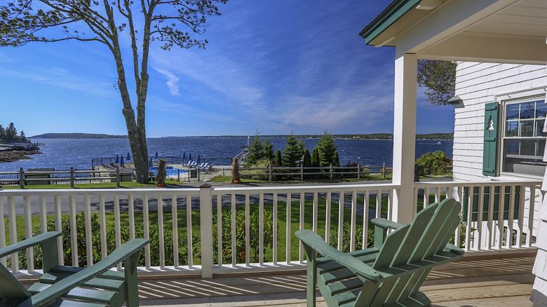 A scenic view from a porch overlooking a body of water, with two green Adirondack chairs, a tree, and a house with white siding and green shutters.