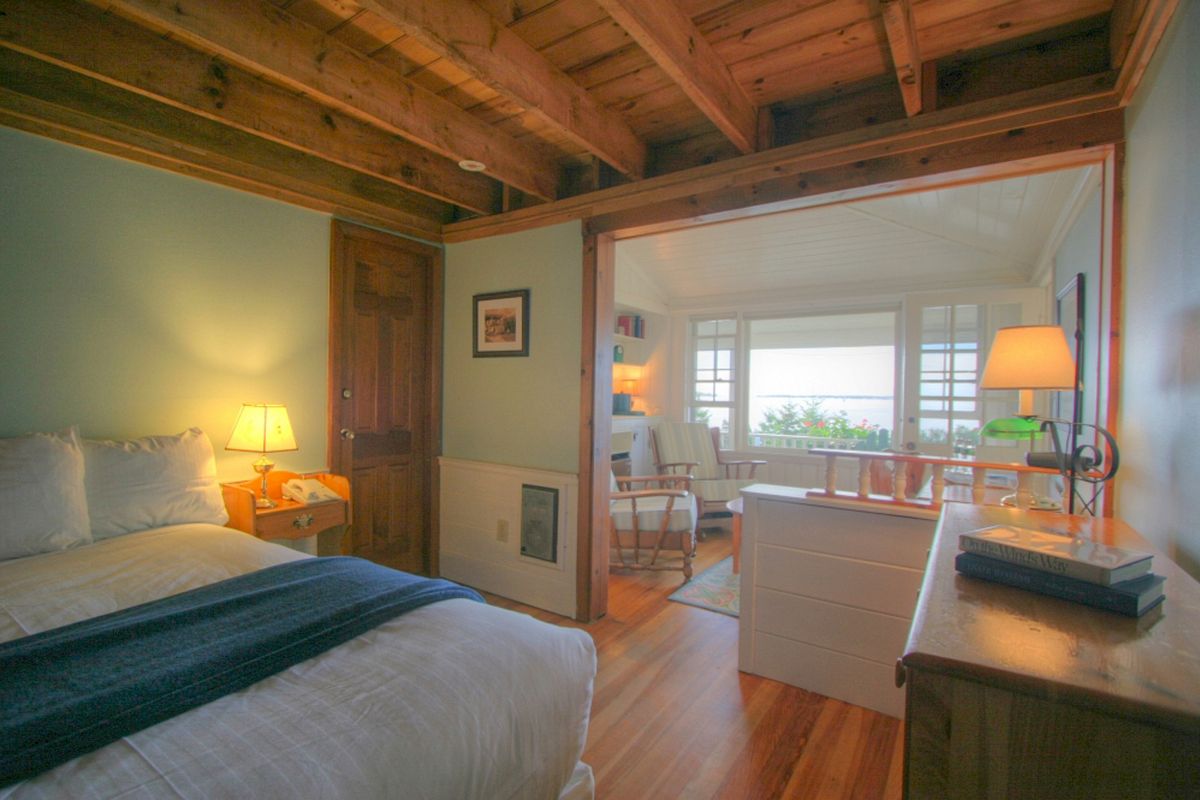 A cozy bedroom with wooden ceiling, bed, lamp, and a view into an adjoining sunroom with more lamps, a chair, and large windows overlooking greenery.