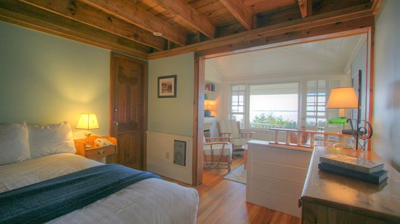 A cozy bedroom with wooden ceiling, bed, lamp, and a view into an adjoining sunroom with more lamps, a chair, and large windows overlooking greenery.