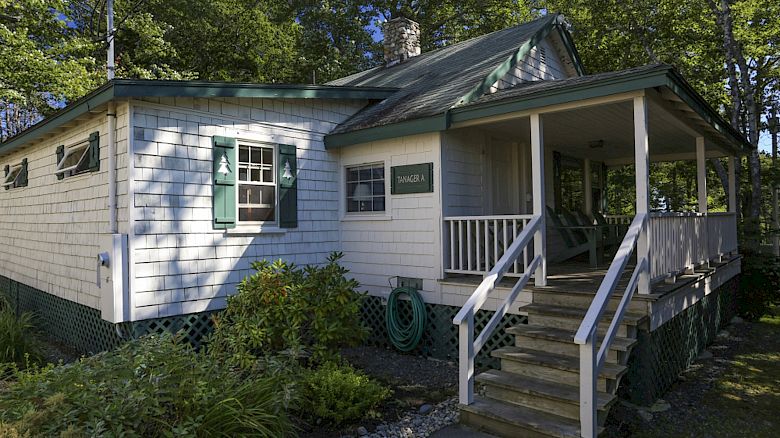 An image of a small house with a shaded porch, green shutters, surrounded by trees and a small garden; there's a hose on the side wall.