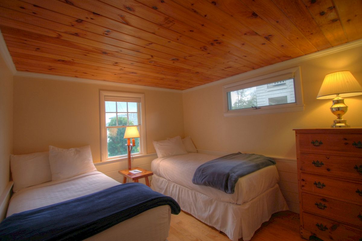 A cozy bedroom with two single beds, a wooden ceiling, a small window, a table lamp, and a dresser with a lamp on top, ending the sentence.