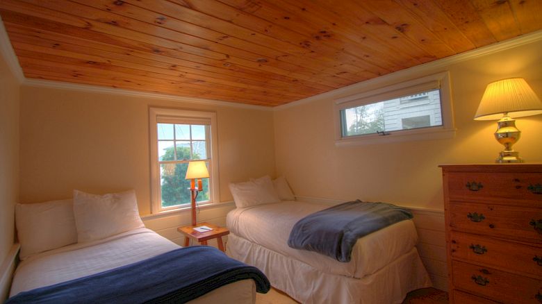 A cozy bedroom with two single beds, a wooden ceiling, a small window, a table lamp, and a dresser with a lamp on top, ending the sentence.