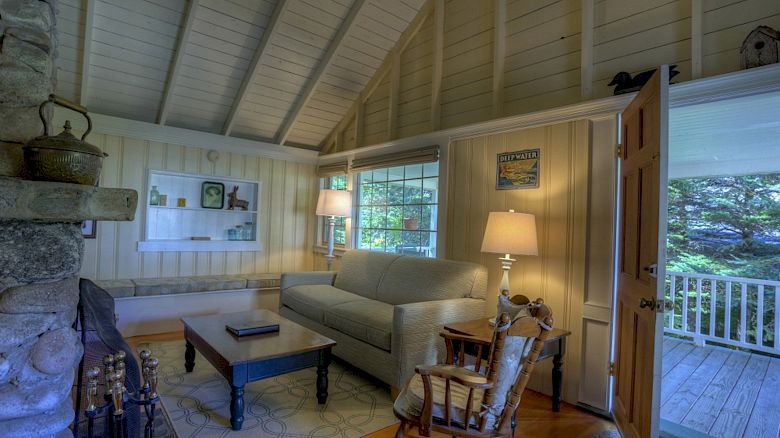 This image shows a cozy living room with a high, vaulted ceiling, a sofa, a coffee table, lamps, and a rocking chair, with an open door leading outside.