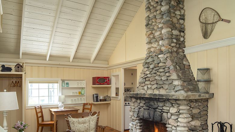 This image shows a cozy living room with a high wooden ceiling, a stone fireplace, a sofa, a coffee table, and a small dining area.