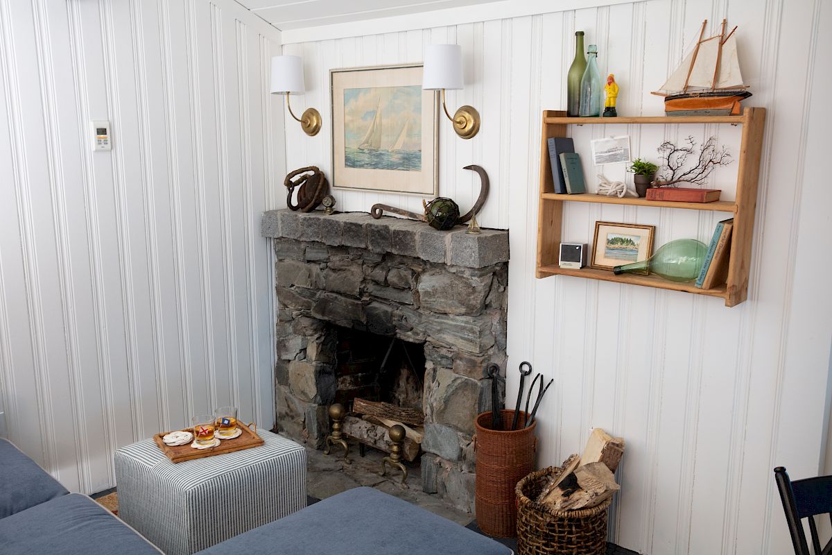 A cozy living room with a stone fireplace, nautical decor, a small ottoman with a tray, and a shelf displaying books, bottles, and small artworks.