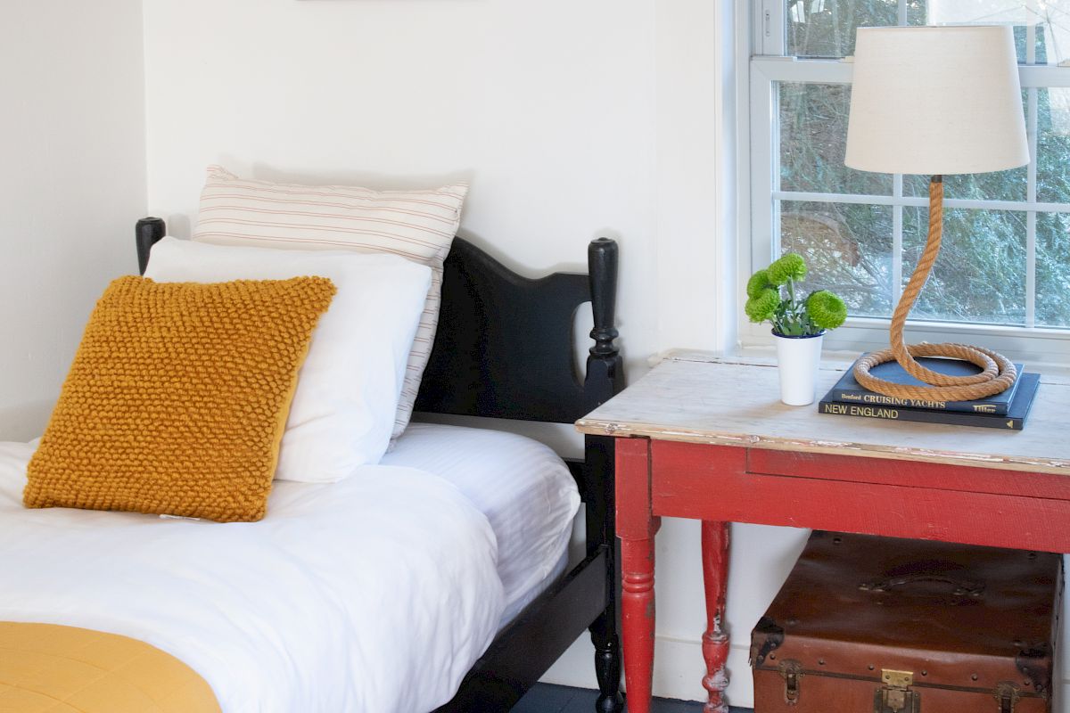 A cozy bedroom features a bed, a bedside table with a lamp, a framed photo on the wall, and a window with woven blinds, ending the sentence.