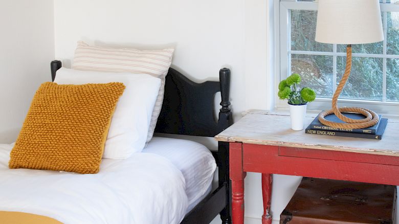 A cozy bedroom features a bed, a bedside table with a lamp, a framed photo on the wall, and a window with woven blinds, ending the sentence.